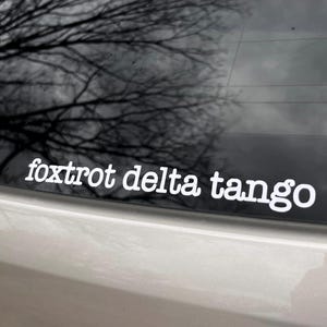 May include: A white vinyl sticker with the text "foxtrot delta tango" on a black car.