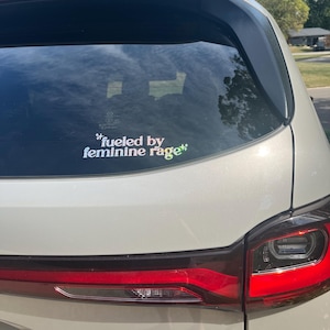 K&ouml;nnte beinhalten: Die Heckscheibe eines wei&szlig;en Autos zeigt einen Aufkleber mit dem Text "fueled by feminine rage" in einer farbenfrohen, stilisierten Schrift. Die R&uuml;cklichter des Autos sind rot, und der Himmel ist durch die Scheibe sichtbar.