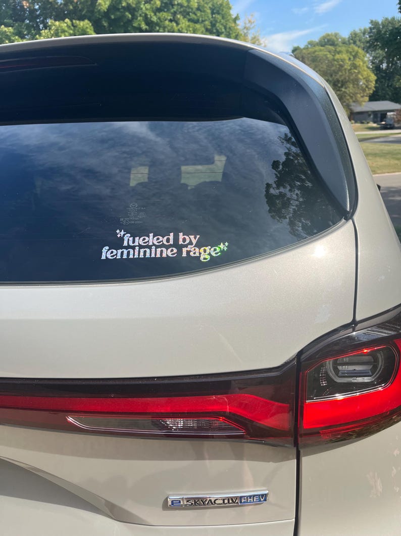 K&ouml;nnte beinhalten: Die Heckscheibe eines wei&szlig;en Autos zeigt einen Aufkleber mit dem Text "fueled by feminine rage" in einer farbenfrohen, stilisierten Schrift. Die R&uuml;cklichter des Autos sind rot, und der Himmel ist durch die Scheibe sichtbar.