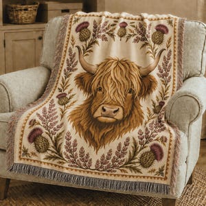 May include: A woven throw blanket featuring a central illustration of a brown Highland cow with long horns, surrounded by a decorative border of purple thistle flowers and green foliage on a cream-colored background with fringed edges.