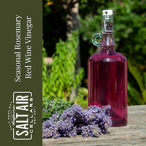 May include: A glass bottle of red wine vinegar with a cork stopper and a label that reads "Salt Air Cellars, California, Half Moon Bay, Seasonal Rosemary Red Wine Vinegar". The bottle is sitting on a wooden surface with lavender flowers and green foliage.