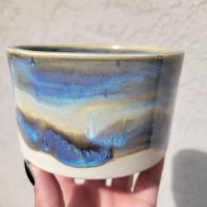 May include: A handmade ceramic bowl with a unique glaze. The bowl features a blend of blue, beige, and brown hues, creating a watercolor effect. The base is a creamy white, and the bowl has a slightly flared shape. The bowl is held in a hand.