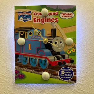 May include: A wall clock featuring a Thomas the Tank Engine design. The clock face is a book cover with the title "Year Round Engines" and the character Thomas. The clock has white, round accents and a gold clock mechanism.
