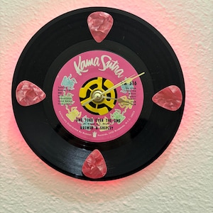 May include: A black vinyl record clock with a pink and yellow label that reads "Kama Sutra" and "One Toke Over the Line." Four pink guitar picks are attached to the record. The clock has a gold-colored clock mechanism.