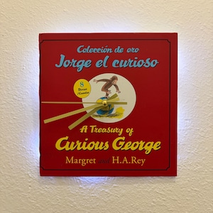 May include: A red square clock featuring the book cover of "A Treasury of Curious George" by Margret and H.A. Rey. The clock face includes the title in yellow and blue, with the Spanish title "Jorge el curioso" at the top. Gold clock hands are visible.