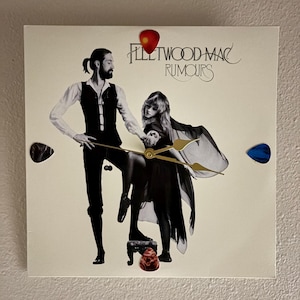 May include: A square clock featuring the Fleetwood Mac Rumours album cover art. The clock face displays the album cover image of two people. The clock hands are gold. Decorative guitar picks in red, brown, and blue are attached to the clock.