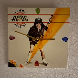 Könnte beinhalten: Quadratische Wanduhr mit dem AC/DC-Logo und dem Albumcover von "High Voltage". Das Zifferblatt zeigt einen Gitarristen mit einem gelben Blitz. Dekorative Gitarrenplektren in verschiedenen Farben sind um das Zifferblatt verstreut.