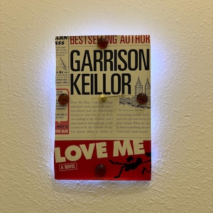May include: A rectangular wall art piece featuring the cover of "Love Me: A Novel" by Garrison Keillor. The artwork includes the book title, author's name, and a red and black illustration. The piece is mounted with decorative red and gold accents.