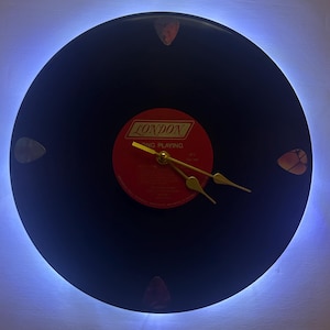 May include: A black vinyl record clock with gold hands and a red label that says "LONDON" and "LONG PLAYING". The clock is surrounded by a blue LED light and has four guitar picks attached to the face.