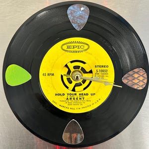 May include: A black vinyl record with a yellow label. The label has the text "EPIC" in a white circle with a red star in the center. The record is titled "Hold Your Head Up" by "Argent". The record is 45 RPM and is a stereo recording. The record is from 1972.