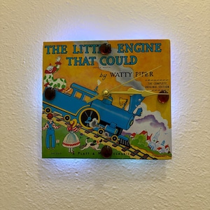 May include: A square clock made from a vintage children's book cover, featuring the title "The Little Engine That Could" and a blue train illustration. The clock has a gold-colored clock hands and four brown decorative accents.