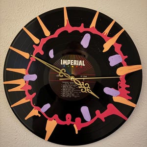 May include: A black vinyl record clock with gold clock hands. The clock face is decorated with orange, pink, and purple paint. The record label reads "IMPERIAL". A unique, handmade wall clock.