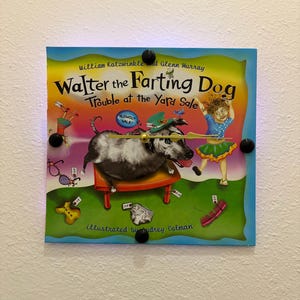 May include: A wall clock featuring the cover of "Walter the Farting Dog: Trouble at the Yard Sale." The book cover art shows a dog on a small table, a girl, and various yard sale items. The clock has black circular accents.