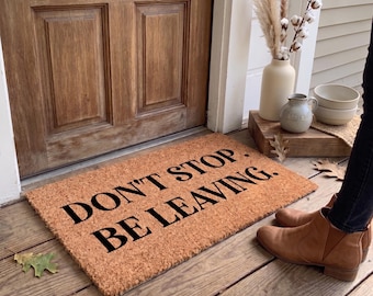 May include: A rectangular coir doormat with the text "DON'T STOP. BE LEAVING." in black. The mat is placed in front of a wooden door. Autumnal decorations, including leaves and a vase, are visible.