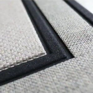 May include: Close-up of a rectangular placemat with a woven texture in shades of grey and beige. The placemat has a black border, creating a defined edge. The design suggests a focus on home decor and dining.