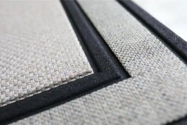 May include: Close-up of a rectangular placemat with a woven texture in shades of grey and beige. The placemat has a black border, creating a defined edge. The design suggests a focus on home decor and dining.