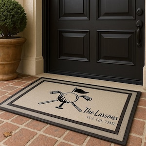 May include: Tan rectangular doormat with a black golf ball, clubs, and flag graphic. The text "The Larsons" and "IT'S TEE TIME" are printed in black. The mat has a dark brown border and is placed in front of a black door.