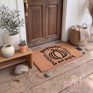 May include: A rectangular doormat with the text "Welcome to our patch" and a pumpkin design in black. The mat is brown and sits in front of a wooden door. A wooden bench with plants and a decorative basket are nearby.