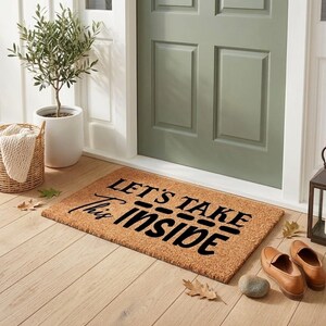 May include: A rectangular doormat with the text "LET'S TAKE THIS INSIDE" in black lettering. The mat is made of a natural fiber material and is placed in front of a green door. A small potted plant, a basket, and a pair of brown shoes are nearby.