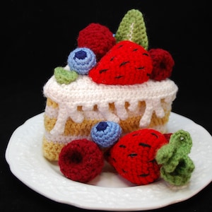 May include: A crocheted cake slice with white icing and yellow cake layers, decorated with red and blue berries and a red strawberry. The cake is on a white plate.