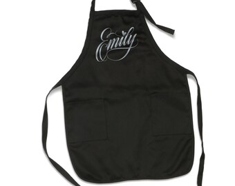 May include: Black apron with two pockets and adjustable straps. The apron has the name "Emily" written in cursive with a heart above the "y".