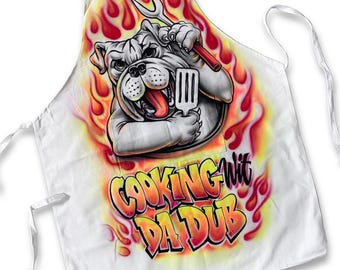 May include: A white apron featuring a graphic of a bulldog holding a spatula and a grilling fork, surrounded by orange and red flames. Below the bulldog, the text "COOKING WIT DA DUB" is printed in a stylized, bold, orange and red gradient font.