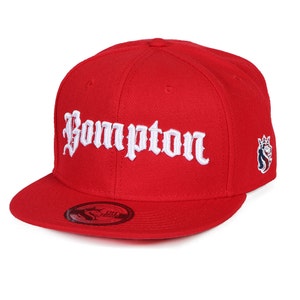 May include: A red baseball cap with a flat brim. The word "Bompton" is embroidered in white script across the front. A small emblem is on the side of the cap.