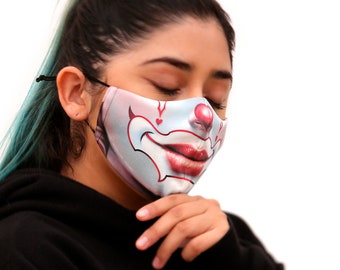 May include: A person wearing a face mask with a clown design. The mask is white with red lips, a red nose, and black eyes. The person has long, dark hair and is wearing a black hoodie.