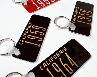 May include: Four vintage-style license plate keychains. Two are black with gold lettering, one says "CALIFORNIA 1959" and the other says "CALIFORNIA 1964". The other two are red and black with white lettering, one says "ARIZONA 1955" and the other says "CALIFORNIA 1958".