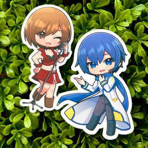 May include: Two anime-style stickers featuring cartoon characters. One sticker shows a brown-haired character in a red outfit with a microphone. The other sticker depicts a blue-haired character in a white and blue outfit.