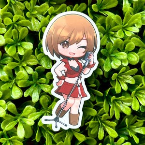 May include: A sticker featuring an anime-style character in a red outfit, holding a microphone. The character has brown hair and is winking. The sticker is set against a background of green foliage.