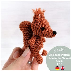 Könnte beinhalten: Gehäkelte Eichhörnchen-Figur in Brauntönen, mit buschigem Schwanz und Haarbüscheln auf dem Kopf. Das Eichhörnchen hat schwarze Augen und einen weißen Fleck auf der Brust. Der Text auf dem Bild lautet "Häkel- Anleitung/Pattern Eichhörnchen/Squirrel Freddy PDF".
