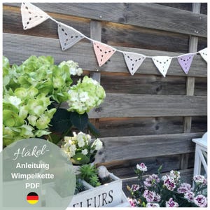 May include: A crochet bunting with pastel pink, white, and purple triangles. The bunting is hanging on a wooden fence. The text "Häkel-Anleitung Wimpelkette PDF" is on a green background with a German flag below the text.