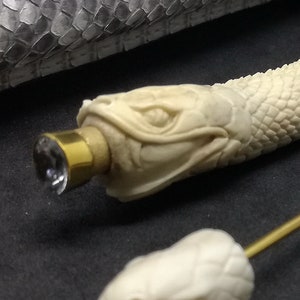 May include: A white bone pen shaped like a snake with a gold-coloured cap and a clear crystal on the end. The pen is lying on a black surface next to a snake-skin patterned case and a gold-coloured metal rod.