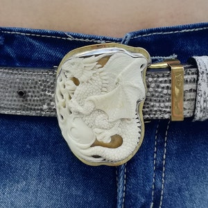 Op de afbeelding: Een witte leren riem met een zilveren gesp met een gegraveerd drakenontwerp. De riem wordt gedragen over een blauwe denim short.