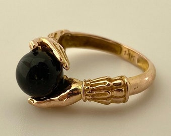 May include: A gold ring with two hands holding a black gemstone. The ring is marked "14K".