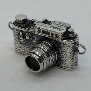 Op de afbeelding: Een gedetailleerde zilveren camera-bedel met een getextureerde afwerking. De camera heeft een lens, zoeker en diverse draaiknoppen, met kleine lussen aan elke kant. Het ontwerp doet denken aan een vintage camera.