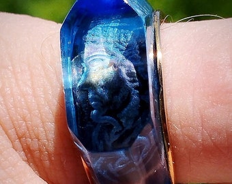 May include: A blue glass ring with a gold band. The ring features a detailed design of a face inside the glass.