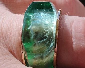 May include: A green glass ring with a gold band. The ring features a cameo design of a woman's face.