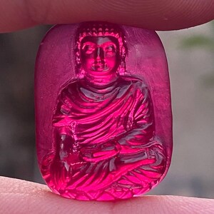 May include: A carved, rectangular, magenta-colored gemstone featuring a seated Buddha figure. The Buddha is depicted in a meditative pose, with detailed folds in the robes. The gemstone is held between two fingers, showcasing its polished surface.