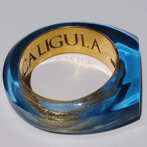 May include: A blue resin ring with a gold interior. The word "CALIGULA" is engraved in the gold band. The ring has a square shape and a translucent appearance.