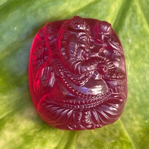 Può includere: Una gemma scolpita rosso rubino raffigurante la divinità indù Ganesha. La figura è seduta, con dettagli intricati nell'intaglio, su uno sfondo di foglie verdi. La gemma è di forma ovale e ha una finitura lucida.