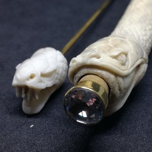 May include: Two intricately carved bone snake head walking sticks with a crystal at the end of one. The snake heads are detailed with scales and fangs.