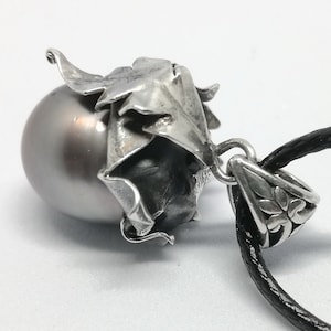 May include: A silver pendant necklace with a large gray pearl and a detailed floral design. The pendant is attached to a black cord.