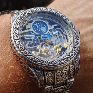 May include: A silver watch with a skeletonized dial and intricate engravings on the case and band. The watch face features a sun and moon design.