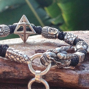 May include: Silver bracelet featuring intricate dragon and geometric designs. The bracelet has a black cord and a series of silver rings hanging from the center. The bracelet is displayed on a wooden surface.