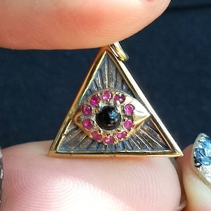 May include: A gold-toned pendant shaped like a triangle with an eye in the center. The eye is surrounded by pink gemstones and a black gemstone in the center. The pendant is being held by a hand with black and white checkered nail polish.