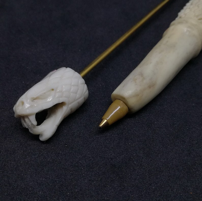 May include: A white bone pen with a snake head design on the top and a gold metal tip. The pen is made of bone and has a natural, organic look.