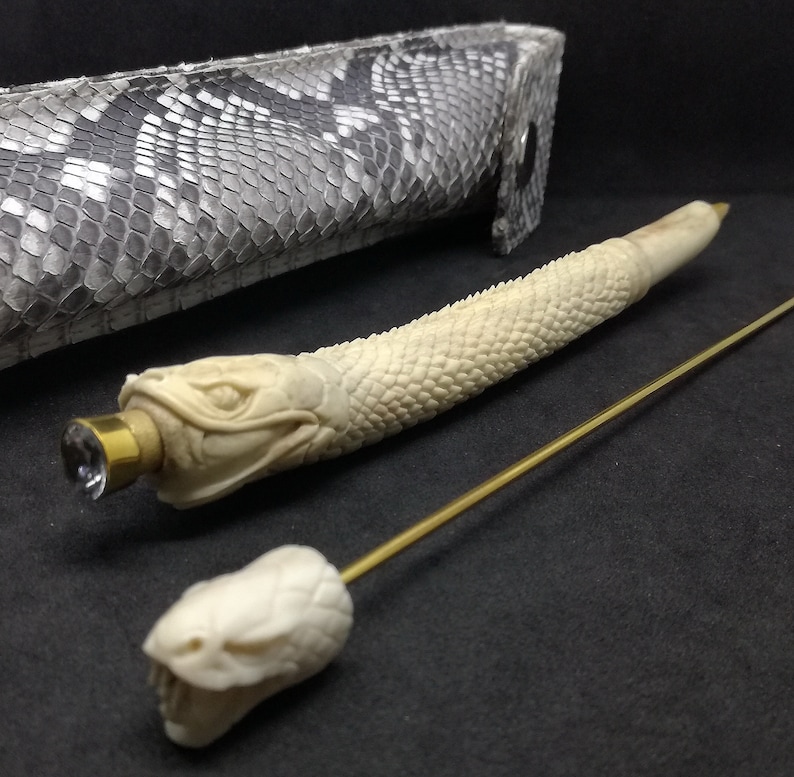 May include: A white bone pen shaped like a snake with a gold-coloured cap and a clear crystal on the end. The pen is lying on a black surface next to a snake-skin patterned case and a gold-coloured metal rod.