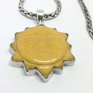 Puede incluir: Un collar de cadena de plata con un colgante de sol amarillo. El sol tiene una cara tallada con una sonrisa.